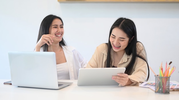 Deux jeunes femmes créatrices de vêtements décontractés riant tout en travaillant avec un ordinateur portable et une tablette numérique dans un bureau moderne.