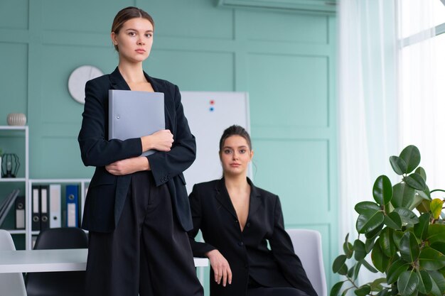 Deux jeunes femmes dirigeantes vêtues d'un costume noir au bureau une femme debout avec un dossier de documents une autre femme assise sur la table regardant la caméra Réunion d'affaires