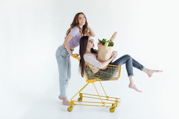 Deux jeunes femmes gaies dans des tenues à la mode colorées souriant et s'amusant avec un chariot