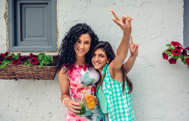 Deux Jeunes Femmes Heureuses Regardant La Caméra Faisant Le Signe De La Victoire Tout En Tenant Des Boissons Saines Sur Fond De Mur