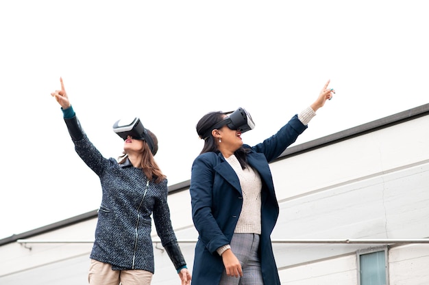 Deux jeunes femmes portant des casques de réalité virtuelle regardent quelque chose et pointent du doigt. Femmes contre un mur de la ville avec espace de copie.