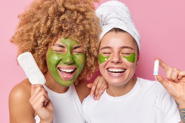 Deux jeunes femmes rient joyeusement subissent des procédures de beauté tiennent la serviette hygiénique et le tampon se tiennent près l'un de l'autre ont une serviette sur la tête isolée sur fond rose Concept d'hygiène et de soins de la peau