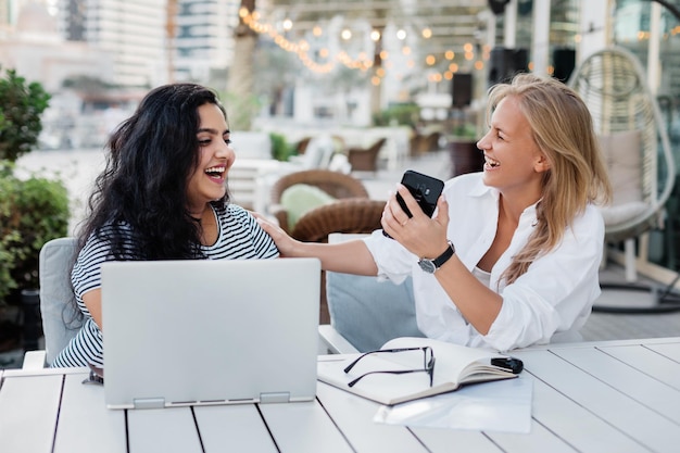 Photo deux jeunes femmes rient pendant une pause dans un café en regardant les matériaux de travail sur un ordinateur portable