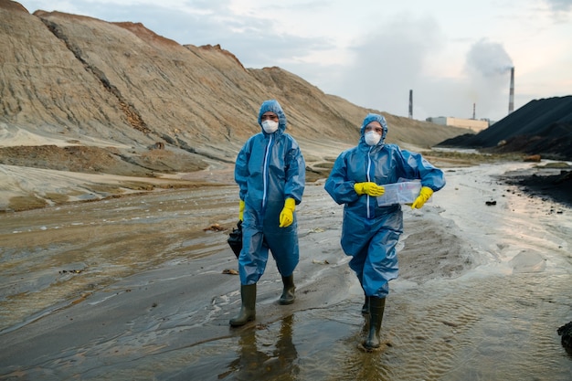 Deux jeunes femmes scientifiques contemporaines portant des combinaisons de protection bleues et des bottes en caoutchouc descendant une rivière polluée entourée de collines