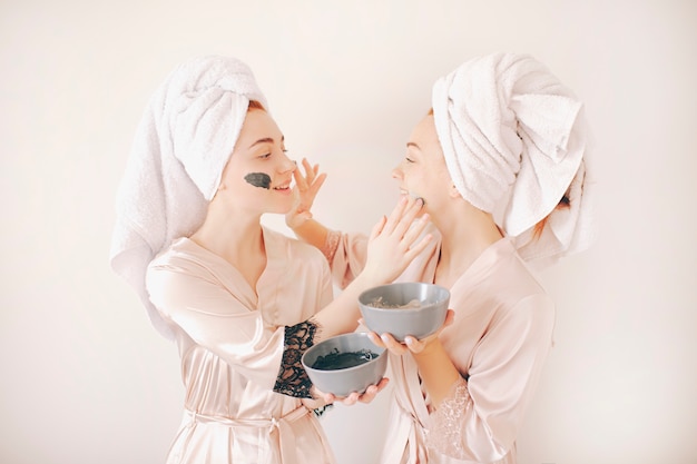 Deux jeunes femmes sœurs en peignoirs avec des serviettes sur la tête isolés sur fond blanc. Spa à domicile. Ils font un masque pour les soins de la peau du visage. Et appliqué sur la peau du visage