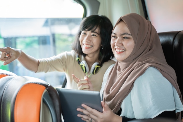 Deux jeunes femmes souriantes avec des doigts pointés tout en discutant tout en tenant une tablette assise sur le bus