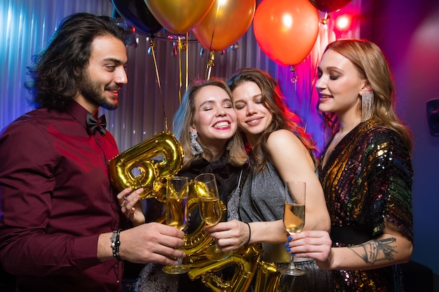 Deux jeunes femmes souriantes tinter des flûtes de champagne avec un homme élégant tout en profitant d'une fête d'anniversaire dans la boîte de nuit