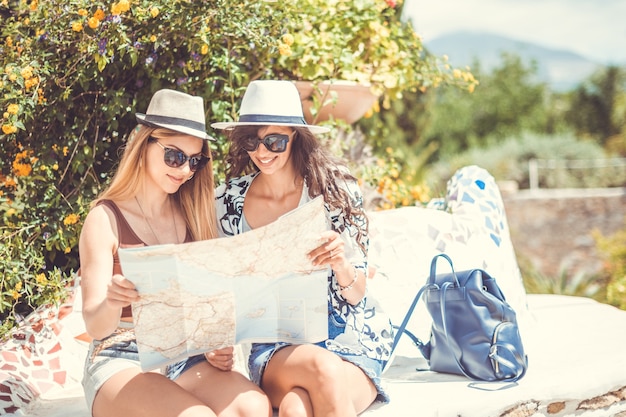 Deux jeunes femmes touristiques souriantes sont assises à la recherche d'une carte pour la direction par une journée ensoleillée