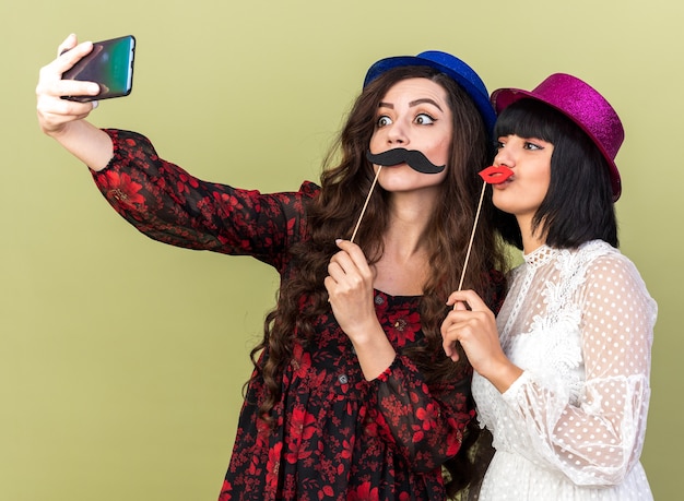 Deux jeunes fêtardes portant un chapeau de fête tenant toutes deux une fausse moustache et des lèvres sur un bâton devant les lèvres prenant un selfie ensemble isolé sur un mur vert olive