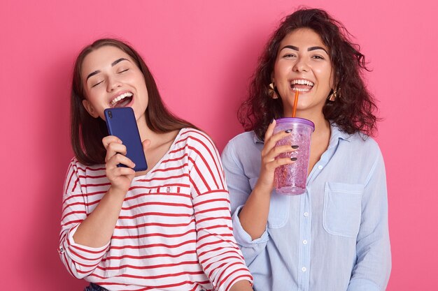 Deux jeunes filles caucasiennes heureuses et excitées chantant ensemble une chanson de karaoké en ligne avec un téléphone et un cocktail comme un microphone