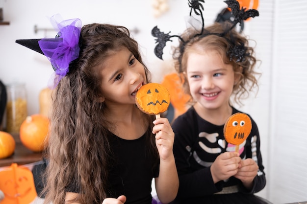 Deux jeunes filles diverses en costume de sorcière, s'amusant dans la cuisine, mangeant des biscuits, célébrant Halloween.