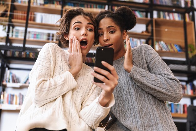 Deux jeunes filles gaies étudiants qui étudient à la bibliothèque, à l'aide d'un téléphone portable, portant des écouteurs