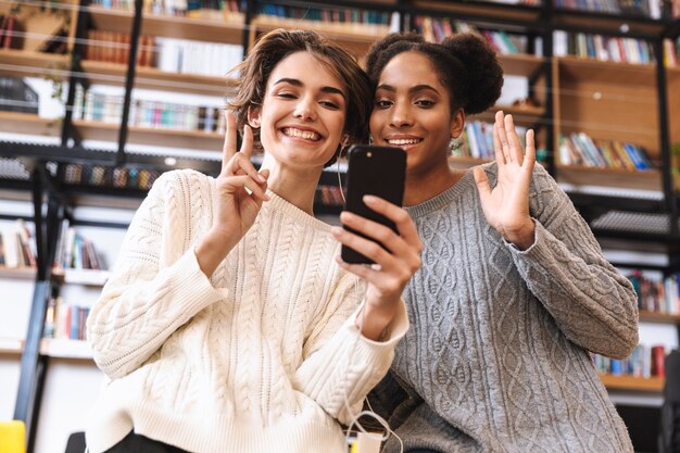 Deux jeunes filles gaies étudiants qui étudient à la bibliothèque, à l'aide d'un téléphone portable, portant des écouteurs