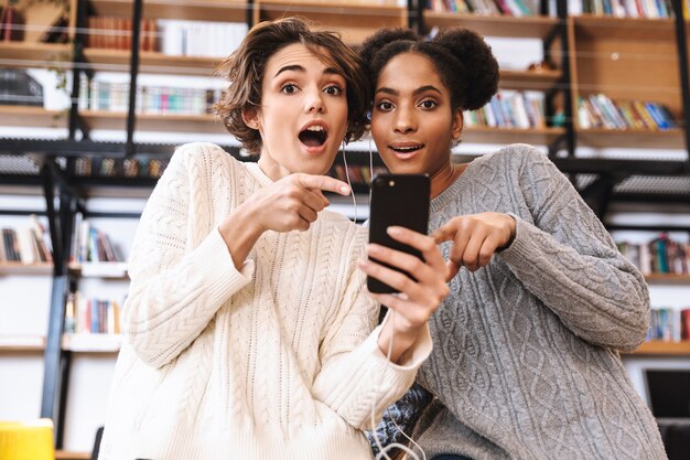 Deux jeunes filles gaies étudiants qui étudient à la bibliothèque, à l'aide d'un téléphone portable, portant des écouteurs