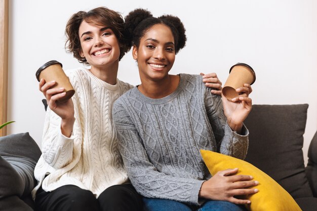 Deux jeunes filles heureux assis sur un canapé ensemble, tenant des tasses à café, tenant des tasses à café