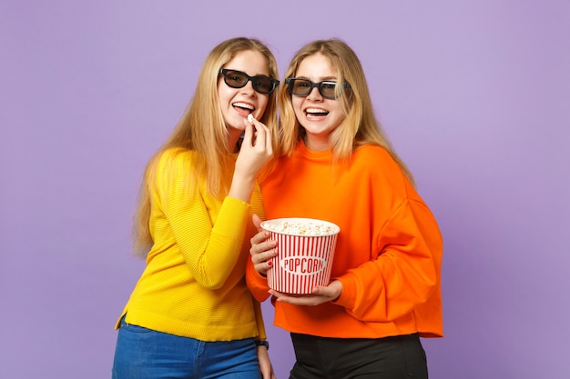 Deux jeunes filles jumelles blondes gaies dans des lunettes imax 3d regardant un film, tenant du pop-corn isolé sur un mur bleu violet pastel. Concept de mode de vie familial de personnes.