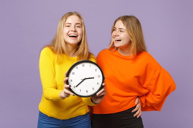 Deux jeunes filles jumelles blondes gaies dans des vêtements colorés vifs tiennent une horloge ronde isolée sur un mur bleu violet pastel. Concept de mode de vie familial de personnes.