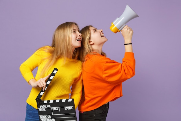 Deux jeunes filles jumelles blondes gaies tiennent un clap de cinéma noir classique, crient sur un mégaphone isolé sur un mur bleu violet. Concept de mode de vie familial de personnes.