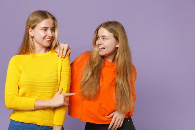 Deux Jeunes Filles Jumelles Blondes Souriantes Dans Des Vêtements Colorés Se Regardant, Pointant L'index Isolé Sur Un Mur Bleu Violet. Concept De Mode De Vie Familial De Personnes.