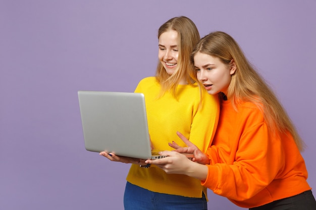 Deux jeunes filles jumelles blondes souriantes dans des vêtements colorés vifs tenant, à l'aide d'un ordinateur portable isolé sur un mur bleu violet pastel. Concept de mode de vie familial de personnes.