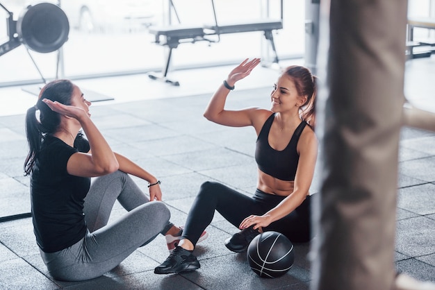 Deux jeunes filles vêtues de vêtements de sport sont ensemble dans la salle de sport pendant la journée, assises sur le sol avec un ballon et donnant un high five.