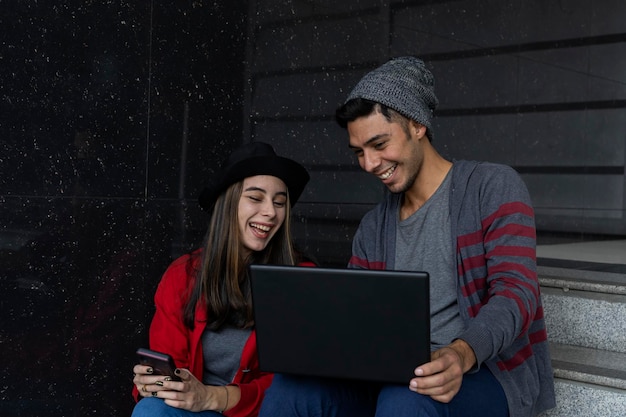 Deux jeunes frères latino-américains assis dans les escaliers avec un ordinateur portable et un téléphone portable tout en partageant du temps de travail en regardant des vidéos et des messages Concept technologique