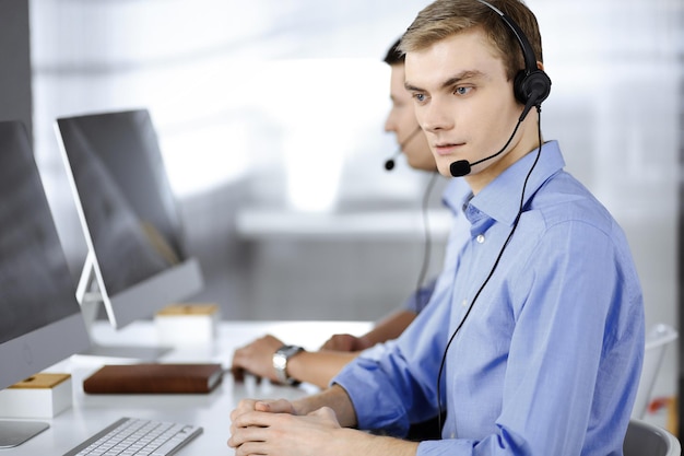 Deux jeunes hommes en casque, assis au bureau dans le bureau moderne, écoutant les clients. Opérateurs de centre d'appels au travail.