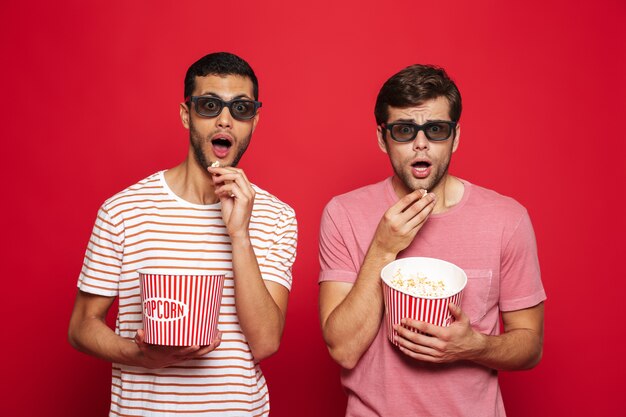 Deux jeunes hommes choqués debout isolé sur mur rouge, manger du pop-corn, portant des lunettes 3d