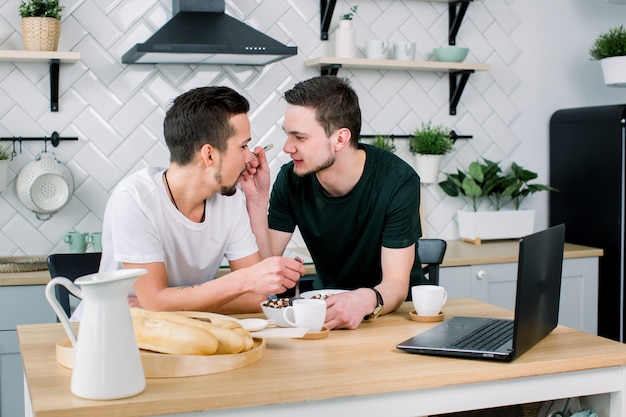 Deux jeunes hommes gais du Caucase heureux se regardant avec des sourires pendant le petit déjeuner. L'homme nourrit son partenaire. Bonjour, concept de couple gay