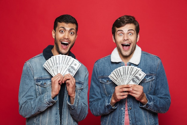Deux jeunes hommes gais isolés sur mur rouge, tenant des billets d'argent