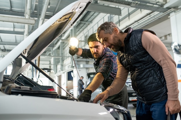 Deux jeunes hommes se penchent sur le compartiment moteur de la voiture ouverte tandis qu'un technicien avec une lampe pointant sur l'une de ses pièces pendant le diagnostic