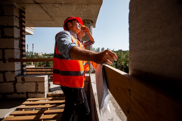 Deux jeunes ingénieurs civils vêtus de gilets de travail orange et de casques se tiennent sur le chantier à l'intérieur du bâtiment en construction .