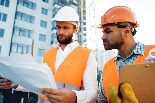 Deux jeunes ingénieurs masculins en uniforme et casques travaillant au chantier de construction