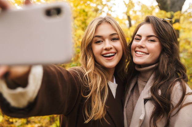 Photo deux jeunes jolies filles gaies portant des manteaux marchant ensemble dans la forêt d'automne, prenant un selfie