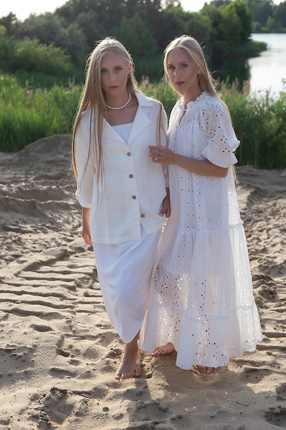 Deux jeunes jolies jumelles aux longs cheveux blonds posant sur la plage de sable en élégante robe blanche