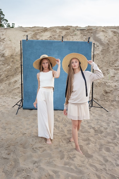 Deux jeunes jolis jumeaux caucasiens aux longs cheveux blonds posant dans la carrière de sable dans d'élégants vêtements blancs, beiges et chapeaux en face de fond bleu