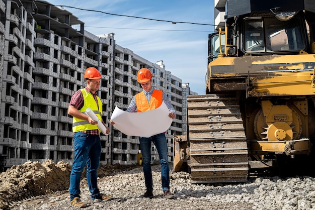 Deux jeunes travailleurs forts en uniforme et casques se tiennent près d'une niveleuse sur un chantier de construction. Concept de construction