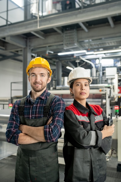 Deux jeunes travailleurs portant des casques et des vêtements de travail croisant les bras par la poitrine en se tenant debout contre l'intérieur du grand atelier de l'usine moderne
