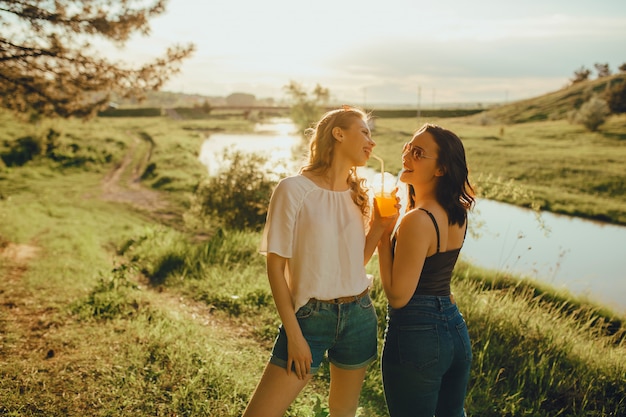 Deux jolies copines boivent du jus d'orange avec de la paille au coucher du soleil, dans des lunettes de soleil, expression faciale positive, en plein air