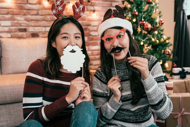 deux jolies dames ludiques positives joyeuses tenant des jouets décorés en papier couvrant les yeux tromper la bonne humeur à l'intérieur ont ouvert la bouche. jeunes amies jouant avec de fausses lunettes moustache à l'arbre de Noël.