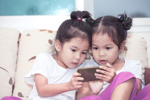 Deux jolies filles d&#39;enfants asiatiques s&#39;amuser à jouer au jeu dans le téléphone intelligent ensemble dans la chambre