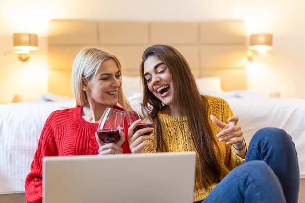 Deux jolies filles joyeuses meilleures amies s'amusant à regarder des films sur un ordinateur portable et à boire du vin rouge à la maison Deux verres de vin blanc dans les mains