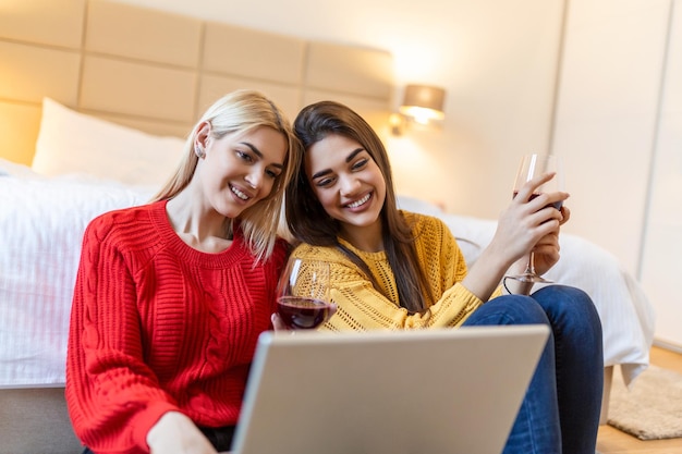 Deux jolies filles joyeuses meilleures amies s'amusant à regarder des films sur un ordinateur portable et à boire du vin rouge à la maison Deux verres de vin blanc dans les mains