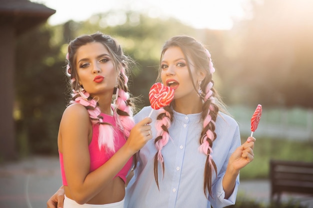 Deux jolies filles à la mode portant des vêtements à la mode avec une tresse colorée et un maquillage professionnel, après le shopping. Femmes marchant ensemble dans la rue, tenant un coeur de bonbons sur un bâton et souriant.