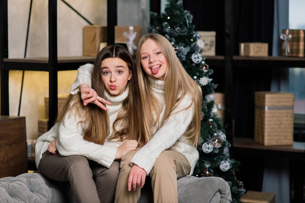 Deux jolies filles posant avec l'expression du visage embrassant avec arbre de Noël. Dame européenne aux cheveux longs debout à côté de l'arbre et des cadeaux