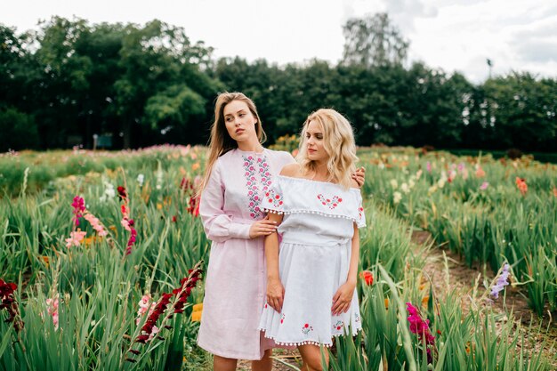 Deux jolies jeunes amies en robes ethniques se détendre dans le jardin fleuri en été. Couple de jolies femmes aux pieds nus dans des broderies de pays se tenant la main et se tenant en plein air. Fille à la mode élégante