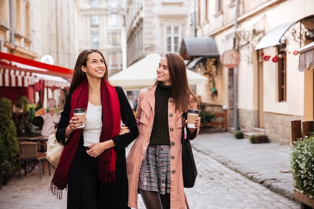 Deux jolies jeunes femmes souriantes marchant et buvant du café dans la vieille ville