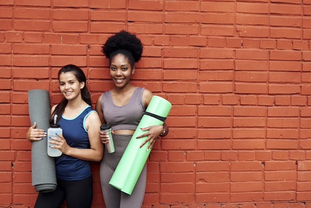 Deux jolies jeunes femmes en survêtement se tiennent contre un mur de briques dans la rue. Les copines sourient tout en tenant des tapis de yoga et des bouteilles d'eau.