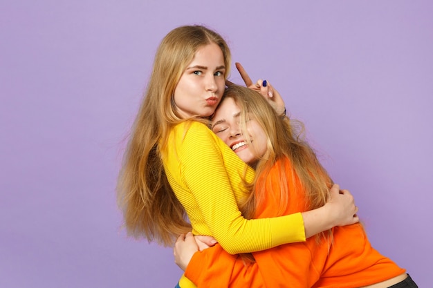 Deux jolies jeunes filles jumelles blondes gaies dans des vêtements colorés vifs étreignant isolés sur un mur bleu violet pastel. Concept de mode de vie familial de personnes.