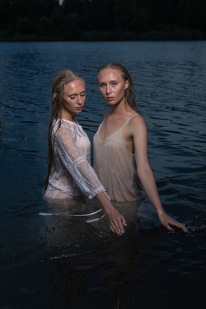 Deux jolies jeunes soeurs jumelles aux longs cheveux blonds posant dans des robes légères dans l'eau du lac pendant la nuit d'été